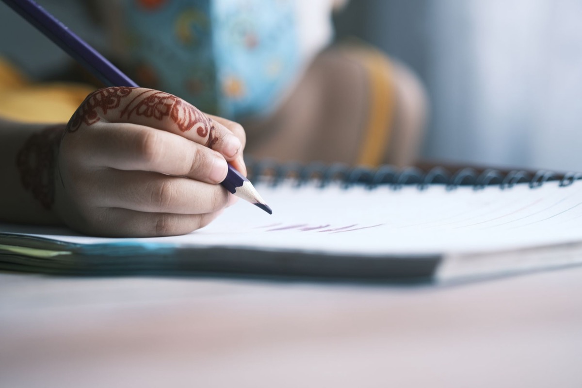A young child practicing handwriting on ruled paper at a school desk, holding a pencil